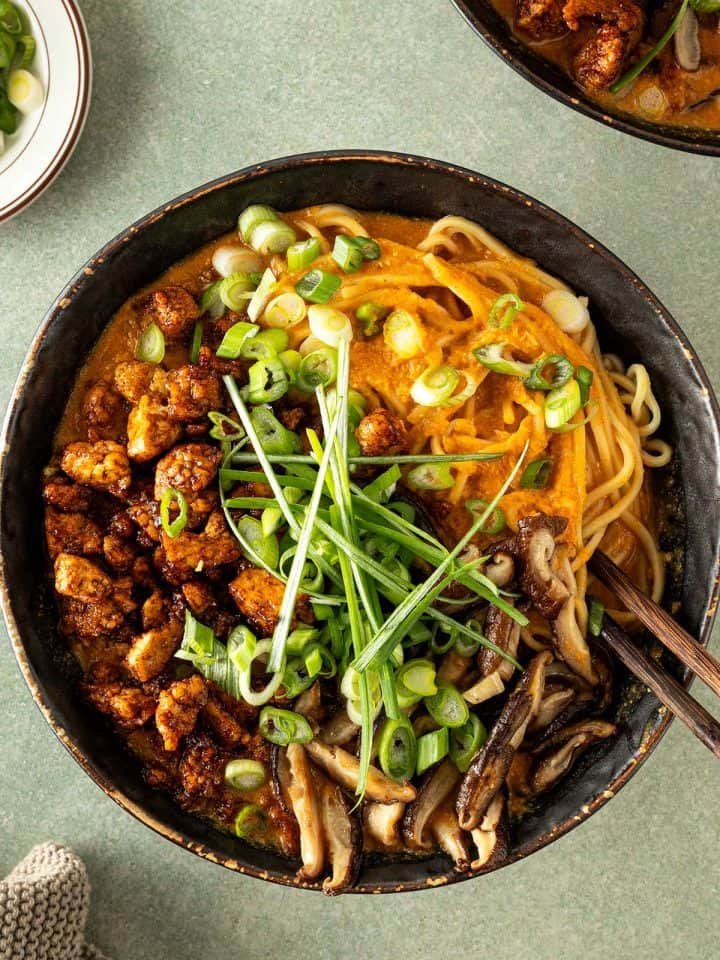 Cremige vegetarische Erdnuss-Ramen mit Ramennudeln, knusprig gebratenem Tofu, Shiitakepilzen und Frühlingszwiebeln in einer dunklen Schüssel von oben fotografiert.
