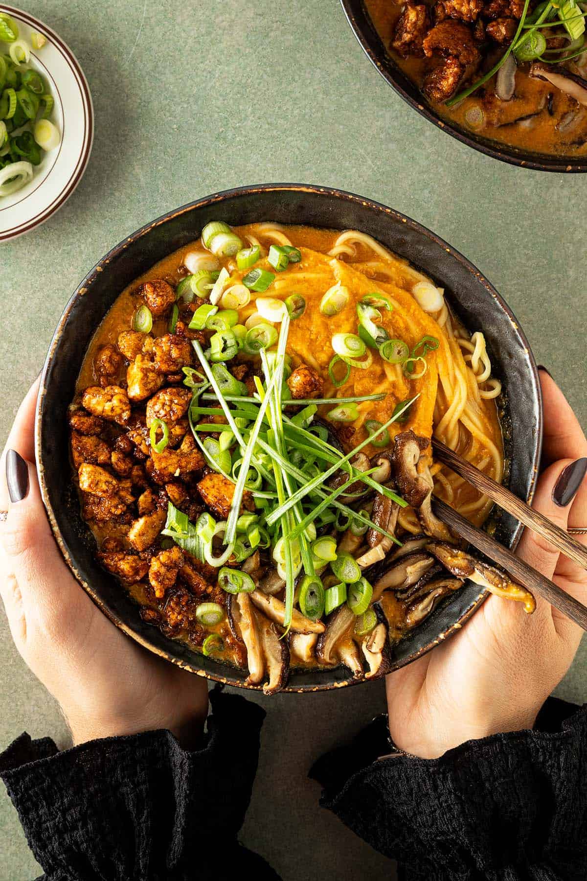 H&auml;nde halten eine Sch&uuml;ssel mit vegetarischen Erdnuss-Ramen mit w&uuml;rzigem Tofu, Shiitakepilzen, Fr&uuml;hlingszwiebeln und cremiger Erdnuss-Miso-Sauce.