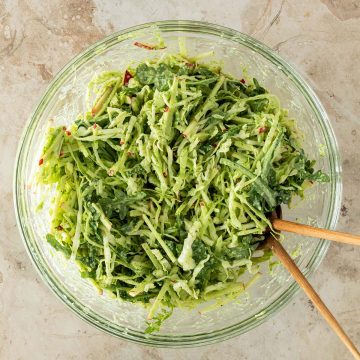 Eine Sch&uuml;ssel mit Wei&szlig;kohlsalat mit Apfel, Rucola und gr&uuml;nem Dressing auf einem Steinuntergrund mit Salatbesteck.