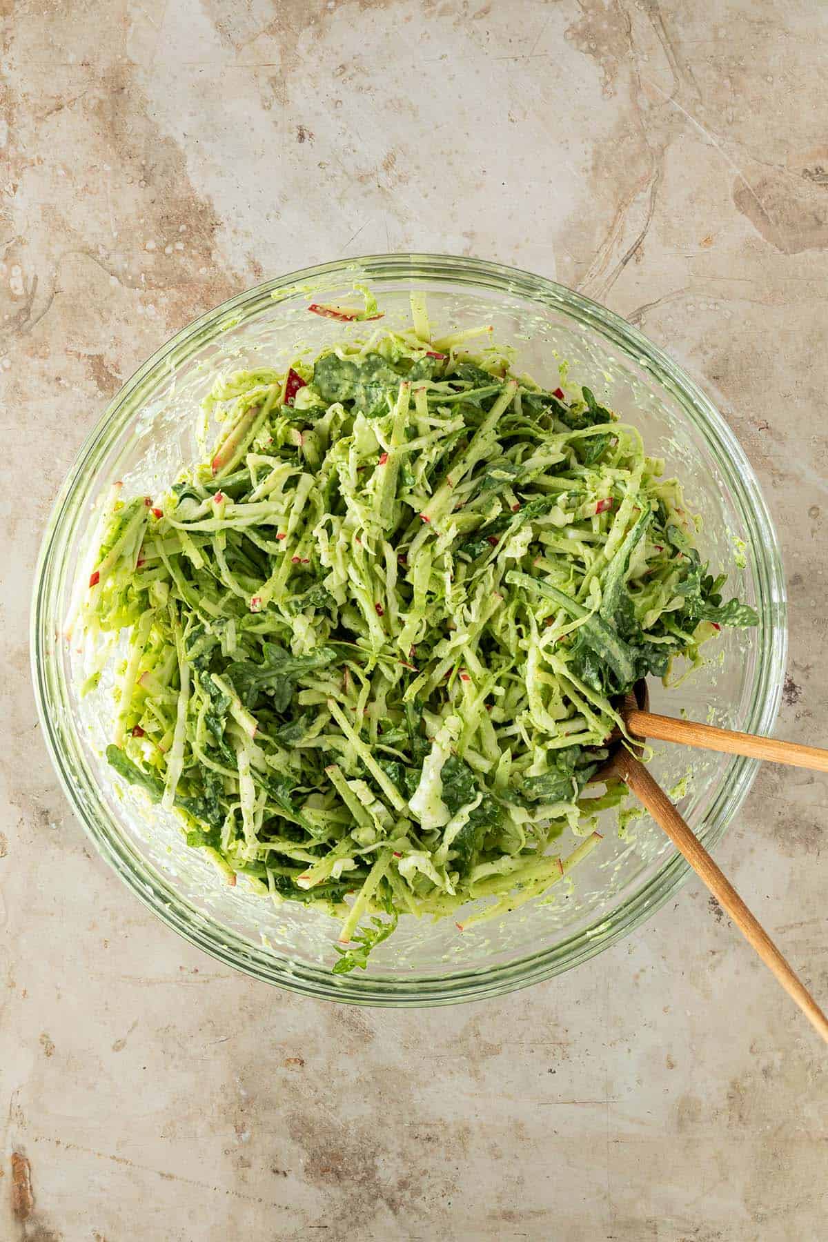 Eine Schüssel mit Weißkohlsalat mit Apfel, Rucola und grünem Dressing auf einem Steinuntergrund mit Salatbesteck.
