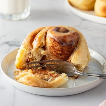 Frische Zimtschnecke mit Zuckerguss auf einem Teller, mit einer Gabel angeschnitten, im Hintergrund ein Glas Milch.