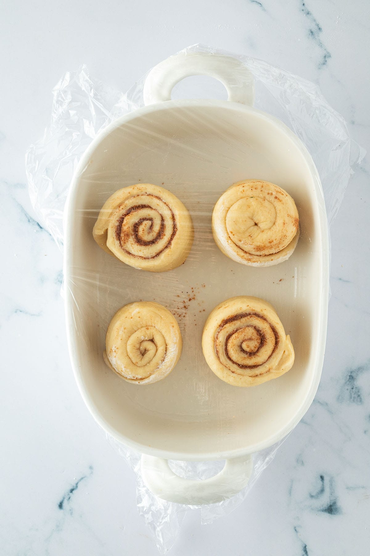 Vier ungebackene Zimtschnecken in einer Auflaufform, abgedeckt mit Frischhaltefolie, bereit zum Aufgehen.
