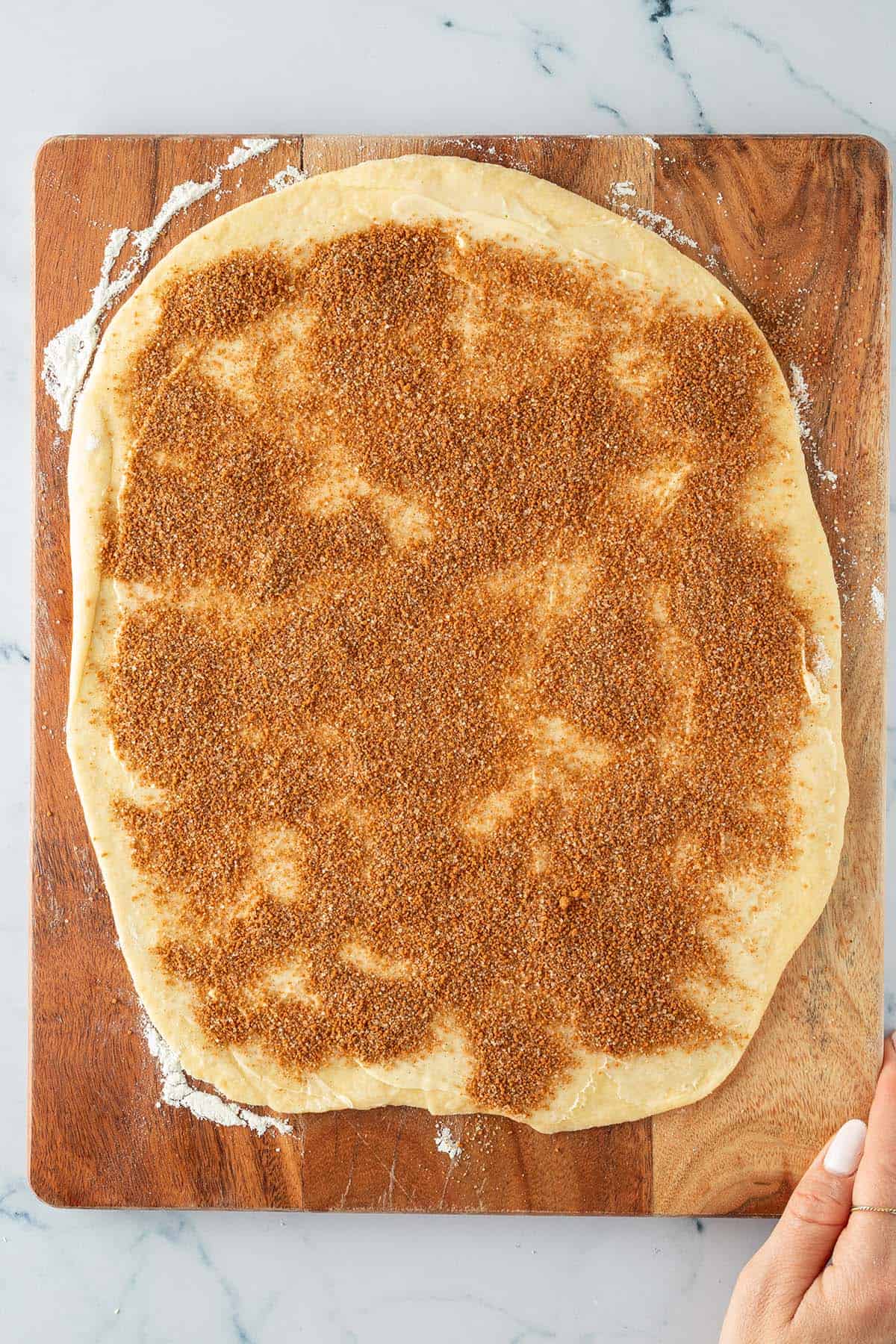 Ausgerollter Hefeteig auf einem Holzbrett, gleichmäßig mit Zimt-Zucker-Mischung bestreut.
