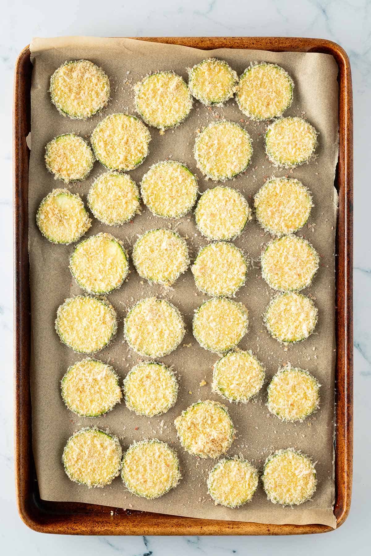 Panierte Zucchini-Scheiben mit Parmesan und Semmelbröseln auf einem Backblech.