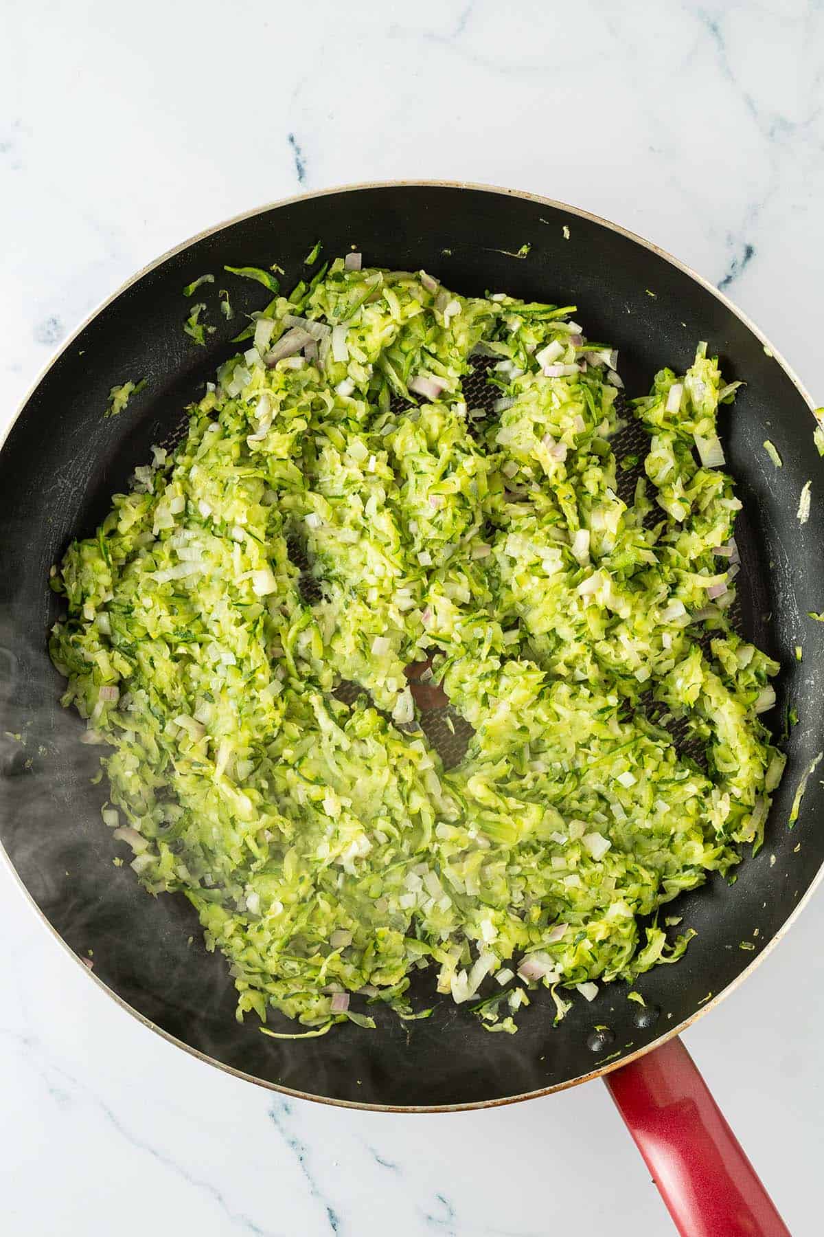 Geraspelte Zucchini und fein gewürfelte Schalotten beim Anbraten in einer Pfanne.